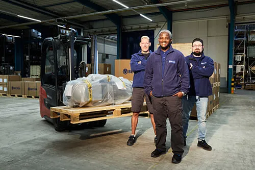 3 Mitarbeiter im Logistikzentrum vor einem Gabelstapler, der eine Palette mit einer Vakuumpumpe angehoben hat