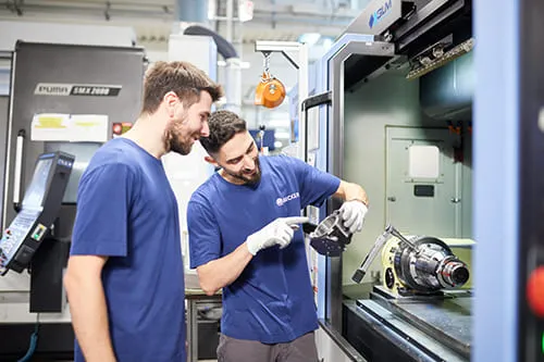 2 Zerspanungsmechaniker begutachten ein Werkstück vor einem CNC-Bearbeitungszentrum