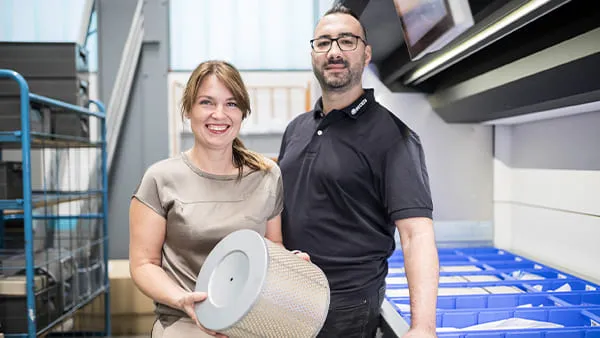 2 Mitarbeitende stehen vor einem Teilelager, eine Person hält eine große Filterpatrone in den Händen
