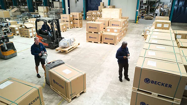 Ein Von-Oben-Ansicht der Versandabteilung, in der an verschiedenen Stellen verpackte Kartons auf Paletten stehen. Logistikmitarbeiter setzen mit Hubwagen und Gabelstaplern Packstücke um